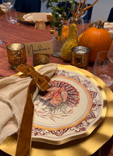 Load image into Gallery viewer, A Turkey & Gourds: Thanksgiving Table for 16 setting featuring a heavy duty paper plate with a turkey illustration, a beige napkin tied with a brown ribbon, and a place card labeled Nandinee, surrounded by gourds and candles on a red tablecloth.