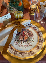 Load image into Gallery viewer, A Sophistiplate LLC Turkey & Gourds: Thanksgiving Table for 16 setting featuring a white turkey-illustrated heavy duty paper plate layered over a gold charger, complete with a cloth napkin, velvet ribbon tie, and a place card labeled Phil.