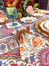 Load image into Gallery viewer, A Turkey & Gourds Guest Towels/20pk harvest pattern napkin decorated with pumpkins and autumn leaves, tucked into a turkey-shaped napkin ring on a patterned tablecloth.