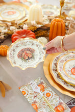 Load image into Gallery viewer, A hand displays the Sophistiplate Turkey & Gourds Table Setting paper plate featuring a turkey design above a festive Thanksgiving table with matching autumn plates, pumpkins, candles, and a patterned napkin.