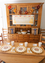 Load image into Gallery viewer, A Thanksgiving table setting featuring the Turkey &amp; Gourds: Thanksgiving Table for 16 set with heavy-duty paper plates, gold chargers, and cups, with a happy fall banner hung on the hutch behind the table.
