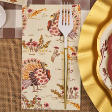 Load image into Gallery viewer, A gold and white fork rests on a Sophistiplate Amber Harvest Guest Towel, decorated with colorful turkeys and flowers, placed on a fall-themed table setting with gold-edged plates and a burlap and plaid tablecloth.
